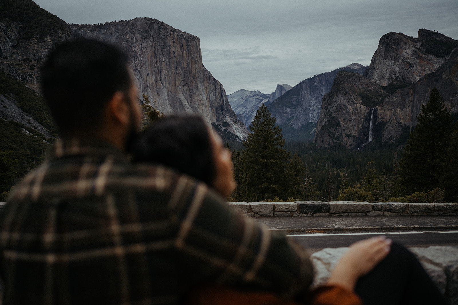 Adventurous couple embracing each other against the breathtaking backdrop of Yosemite National Park. The iconic Tunnel View frames the scene, showcasing majestic granite cliffs and towering trees on a gloomy yet atmospheric day.