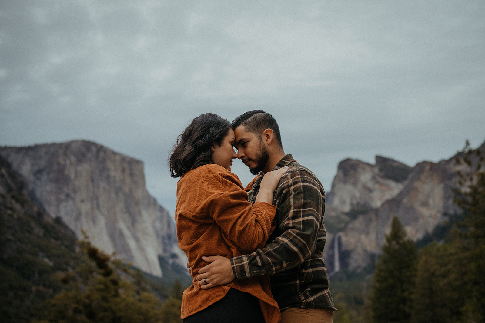 Adventurous couple embracing each other against the breathtaking backdrop of Yosemite National Park. The iconic Tunnel View frames the scene, showcasing majestic granite cliffs and towering trees on a gloomy yet atmospheric day.