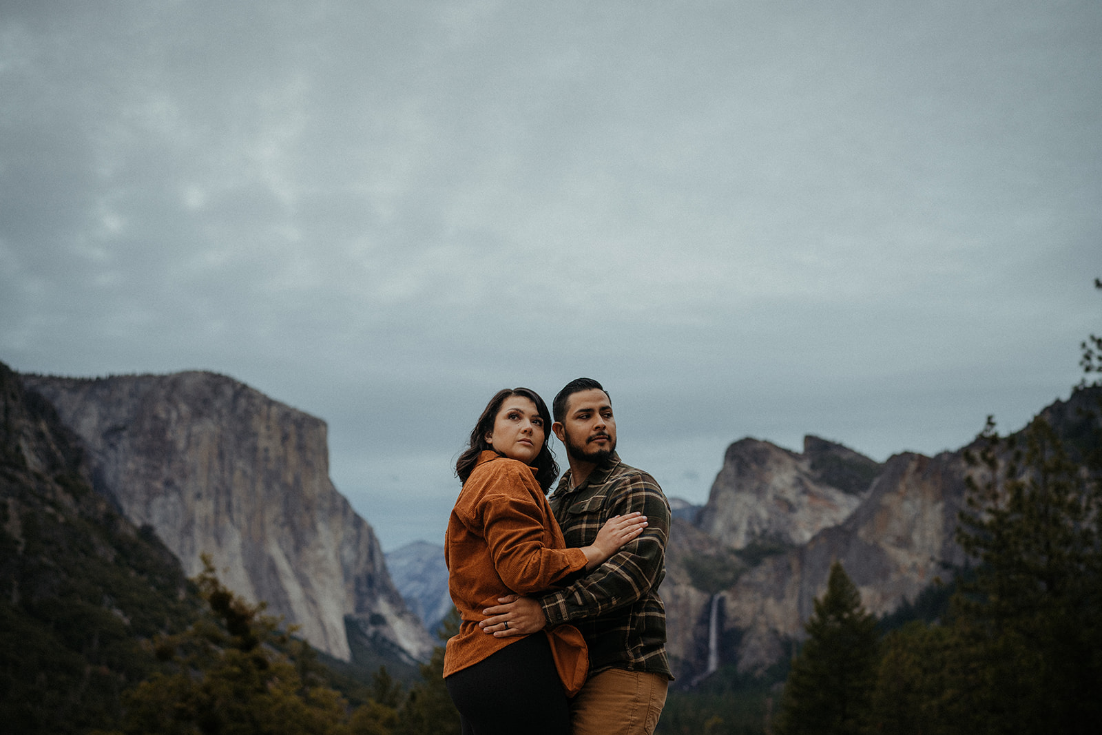 Adventurous couple embracing each other against the breathtaking backdrop of Yosemite National Park. The iconic Tunnel View frames the scene, showcasing majestic granite cliffs and towering trees on a gloomy yet atmospheric day.