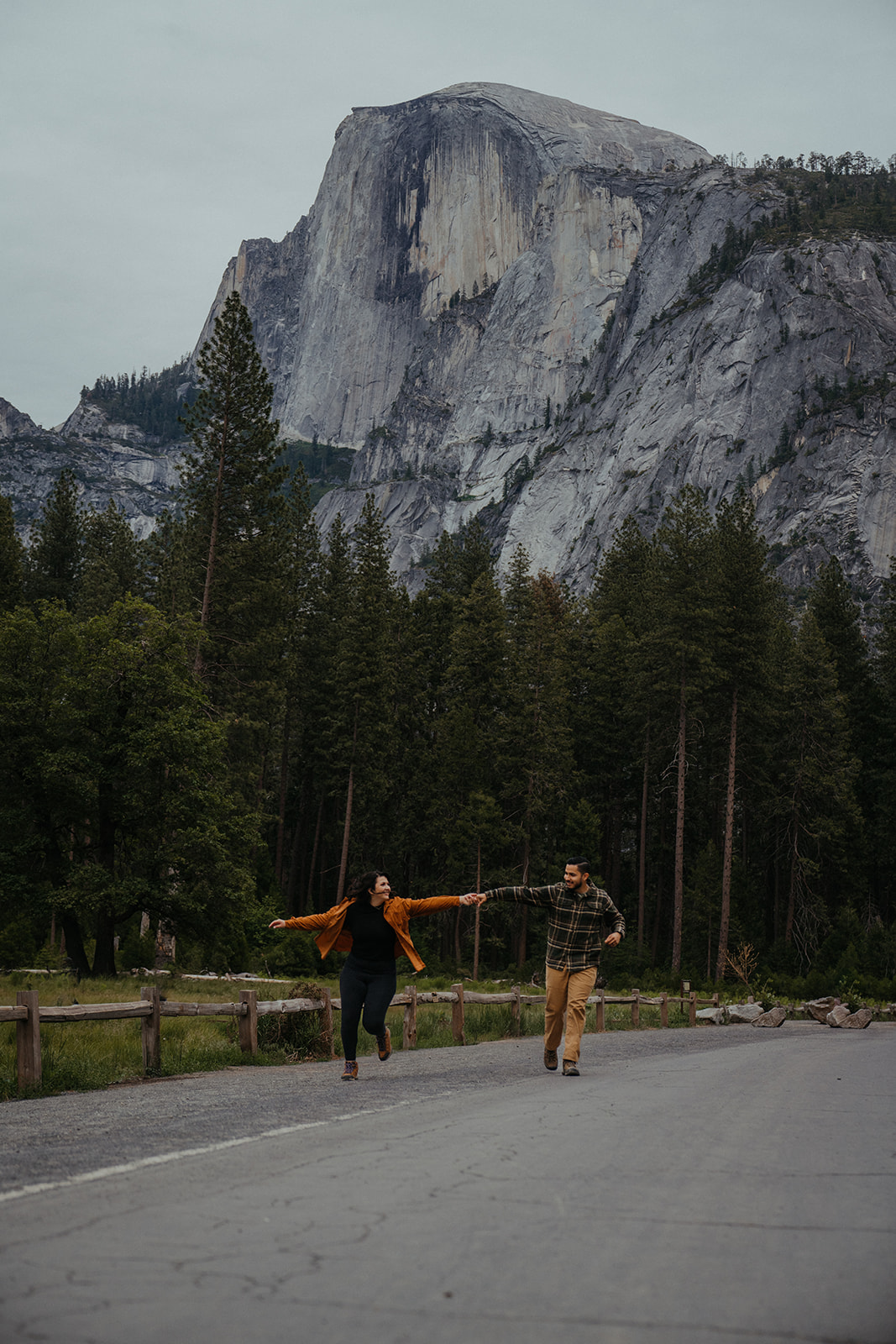 Adventurous Couple runs through the street on the Yosemite Valley floor, holding hands with Half Dome in the background